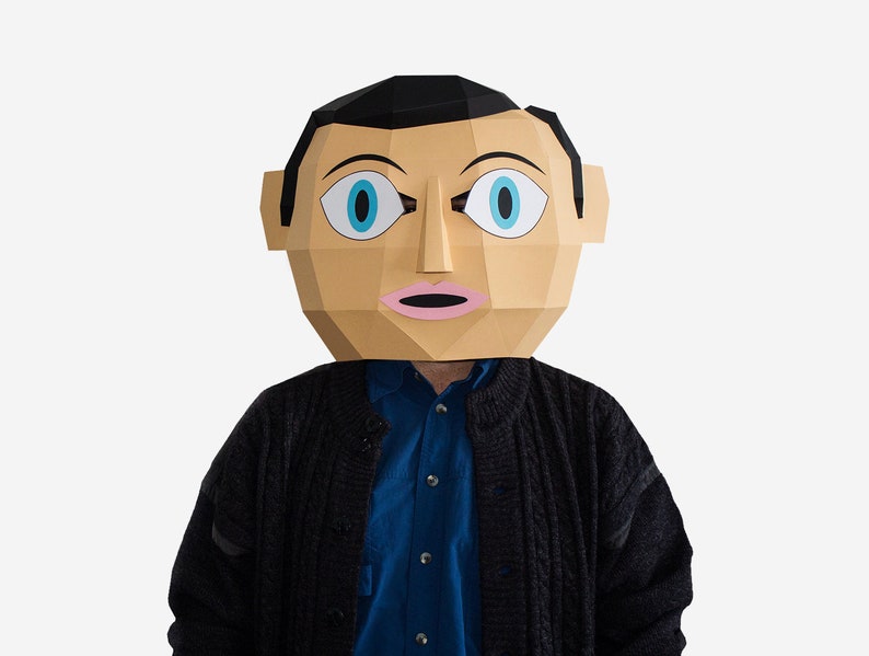 May include: A person wearing a paper mache mask with a surprised expression. The mask is brown with blue eyes and pink lips. The person is wearing a black sweater over a blue shirt.