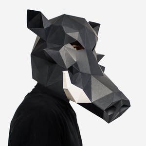 May include: A black paper mache mask of a wild boar with white tusks. The mask is designed with geometric shapes and is worn by a person.