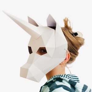 May include: A white paper unicorn mask with a geometric design. The mask is being worn by a person with brown hair.