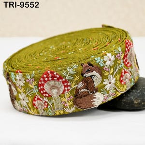 May include: A roll of olive green embroidered ribbon featuring a woodland design. The design includes a brown fox, red and white mushrooms, and white flowers with green leaves. The text "TRI-9552" is visible on the top left.