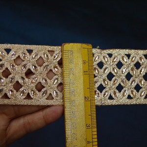 May include: A gold-toned decorative trim featuring a repeating lattice pattern embedded with small, clear, oval-shaped crystals. A yellow wooden ruler is placed vertically against the trim, showing a width of approximately 1.5 inches.