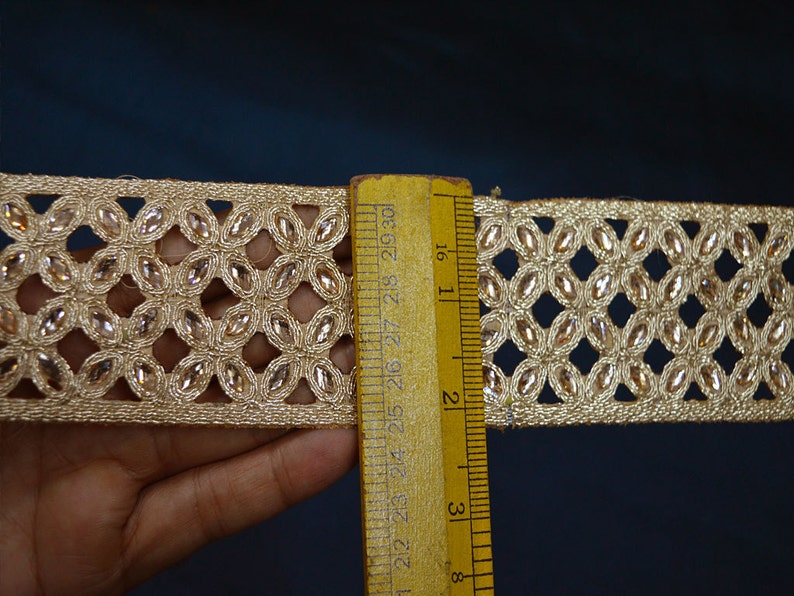 May include: A gold-toned decorative trim featuring a repeating lattice pattern embedded with small, clear, oval-shaped crystals. A yellow wooden ruler is placed vertically against the trim, showing a width of approximately 1.5 inches.