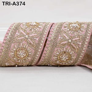May include: A pink fabric ribbon labeled TRI-A374, featuring intricate gold metallic embroidery in floral and leaf patterns, accented with small white beads and circular motifs arranged in a repeating decorative border design.