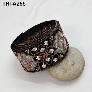 May include: A roll of black embroidered trim featuring floral and leaf designs in white, gold, and pink. The trim is displayed on a smooth, grey stone. The text "TRI-A255" is visible.