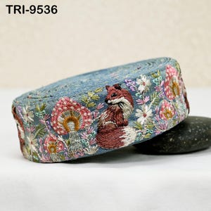 May include: A roll of blue fabric trim featuring intricate embroidery of a brown fox, pink and orange mushrooms, and white flowers. The text TRI-9536 is visible in the upper left corner. The trim is coiled and displays a repeating nature-themed pattern.