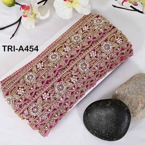 May include: A magenta fabric trim featuring intricate gold embroidery, floral patterns, and clear circular embellishments. The trim is labeled with the text TRI-A454. The design includes repeating circular motifs surrounded by gold thread leaves.