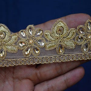 May include: A gold embroidered lace trim featuring a repeating floral pattern with metallic gold thread and clear, teardrop-shaped crystals set within the petals. The design is stitched onto a sheer, light beige mesh fabric with a scalloped edge.