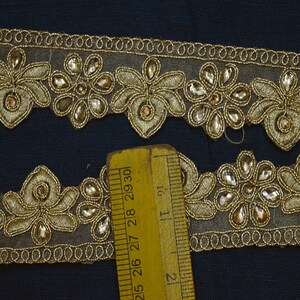 May include: A gold embroidered trim featuring floral motifs and clear teardrop-shaped stones on a sheer mesh base. A yellow ruler is positioned over the trim, showing a width measurement of approximately 1.25 inches for the decorative section.