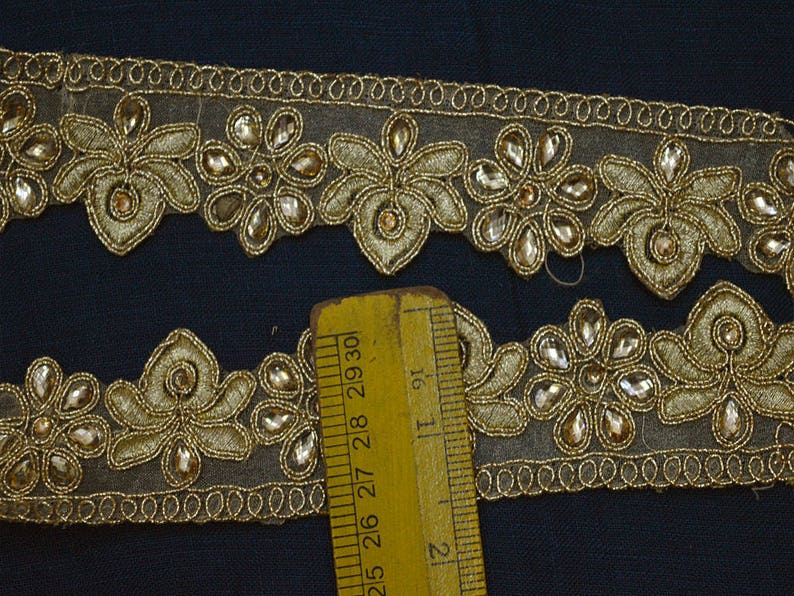 May include: A gold embroidered trim featuring floral motifs and clear teardrop-shaped stones on a sheer mesh base. A yellow ruler is positioned over the trim, showing a width measurement of approximately 1.25 inches for the decorative section.