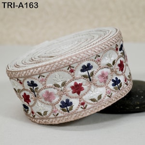 Pode incluir: Um rolo de acabamento bordado branco com um padrão floral. O acabamento apresenta um design repetido de flores vermelhas, rosa e azuis com folhas verdes. As bordas são aparadas com uma borda marrom clara. O texto "TRI-A163" é visível.