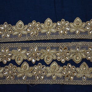 May include: Three strips of gold-toned decorative trim featuring a repeating floral pattern. Each flower is crafted with metallic gold thread embroidery and accented with clear, teardrop-shaped crystals set within the petals on a sheer mesh base.