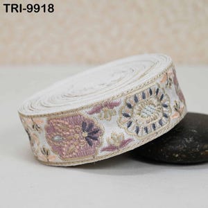 May include: A roll of white embroidered ribbon with a floral design in shades of pink, blue, and peach. The ribbon has a cream-colored border and the text "TRI-9918" is visible.