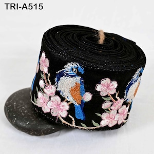 May include: A roll of black fabric ribbon featuring embroidered blue birds and pink cherry blossoms. The text TRI-A515 is printed in the upper left corner. The ribbon is coiled and displays a repeating pattern of birds perched on branches.