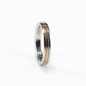 May include: A silver-colored ring with a gold-colored band in the center. The gold band has a textured, ridged design. The ring is set against a white background, highlighting its metallic sheen and simple elegance.