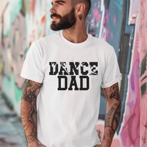 May include: White t-shirt with the text "DANCE DAD" in black block letters. The word "DANCE" features silhouettes of dancers. The shirt is short-sleeved and the person wearing it has tattoos on their arm.