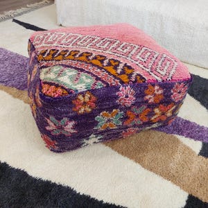 May include: A square pouffe with a colourful, patterned design. The top is pink with a geometric pattern, transitioning to orange and purple floral patterns on the sides. The pouffe sits on a patterned rug.