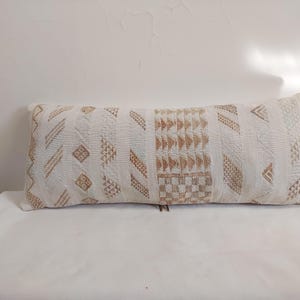 Peut inclure: Long coussin rectangulaire avec un fond blanc et des motifs géométriques beiges et bruns. Les motifs comprennent des rayures, des losanges et des triangles. Le coussin mesure environ 60 cm de long et convient à un canapé ou un lit.
