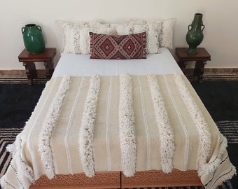 May include: A bed is made with a white sheet, adorned with cream-colored pillows and a long, cream-colored blanket with vertical fringed accents. A patterned, burgundy pillow sits in the center. Two green vases sit on wooden side tables.