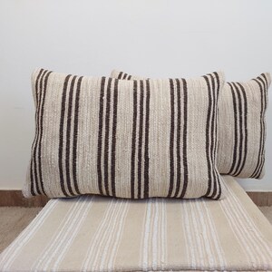 Peut inclure: Deux coussins rectangulaires beiges à rayures verticales marron foncé. Les coussins sont placés sur un coussin beige à rayures. Le fond est un mur blanc uni.