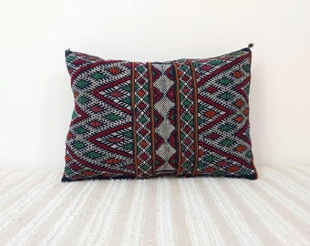 May include: A rectangular pillow with a geometric pattern in shades of red, green, blue, and white. The pillow has a black border and is made of a woven fabric.