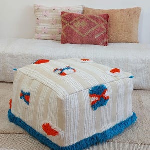 May include: A cream-coloured, striped pouffe with colourful accents. The square pouffe has orange, blue, and white patches. It features a bright blue fringe around the base. Decorative pillows in pink and peach tones are in the background.