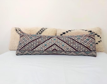 May include: A long, rectangular pillow with a geometric pattern in shades of blue, red, and white. The pillow is made of a woven fabric and has a fringe border. The pillow is sitting on a white bedspread.
