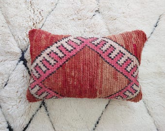 Può includere: Un cuscino rettangolare tessuto a mano con un design geometrico rosso e rosa. Il cuscino ha una superficie strutturata ed è posto su un tappeto bianco e soffice. Il design del cuscino presenta un motivo a diamante centrale.