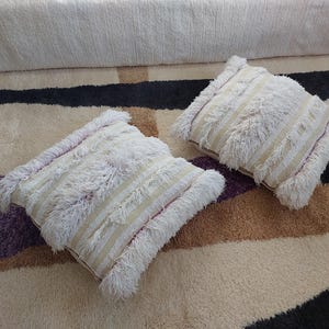 Peut inclure: Deux coussins décoratifs carrés blanc cassé avec une texture hirsute et frangée. Les coussins ont des rayures verticales de couleur beige clair. Les coussins sont sur un tapis à motifs avec des sections beiges, marron, noires et violettes.