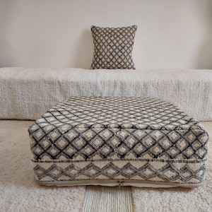 May include: A square, cream-colored ottoman with a black diamond pattern. A matching pillow sits on a white textured surface. The ottoman is a low, floor-level seating option, perfect for a living room or bedroom.