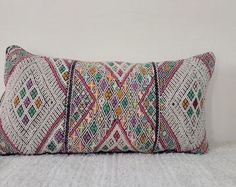 May include: A long rectangular pillow with a white background and a colorful geometric pattern in shades of pink, green, purple, and orange. The pattern is made up of diamonds and triangles.