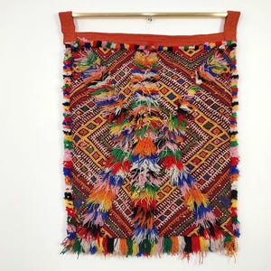 May include: A colorful woven wall tapestry with a geometric pattern and fringe. The tapestry is made of red, orange, yellow, green, blue, and purple yarn.