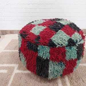 Peut inclure: Un pouf rond avec un motif géométrique rouge, noir et turquoise. Le pouf a une texture touffue et est placé sur un tapis à motifs. C'est un élément décoratif.
