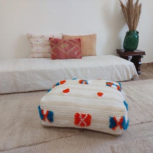 May include: A cream-colored, square pouf with orange and blue accents. The pouf is placed on a neutral-toned rug, with a daybed and pillows in the background. The room has a neutral color palette, with a green vase and wooden side table.