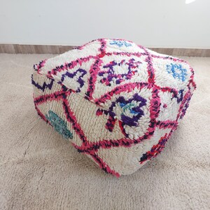 Puede incluir: Puf marroquí cuadrado blanco con un patrón geométrico en rosa, morado y azul. El puf está hecho de una tela texturizada y se asienta sobre una alfombra de color claro. El diseño presenta un patrón de cuadrícula con acentos florales.