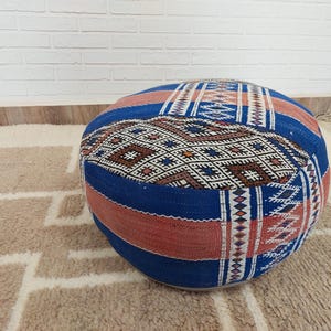 May include: Round, patterned floor pouf in shades of blue, red, and brown. The pouf features a geometric design with a mix of colors and textures. It is placed on a patterned rug, adding a touch of bohemian style to any space.