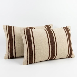 Peut inclure: Deux coussins rectangulaires présentant une texture tissée de couleur beige clair. Chaque coussin est décoré de trois bandes verticales distinctes de couleur brun foncé qui parcourent la surface avant, créant un motif uniforme sur les deux.