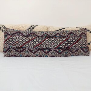 Puede incluir: Una almohada larga y rectangular con un patrón geométrico en tonos rojos, azules y blancos. La almohada está hecha de una tela tejida y tiene un borde de flecos.