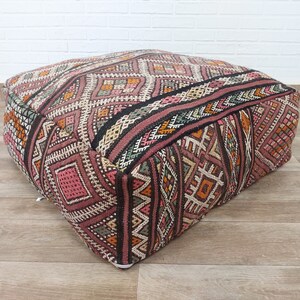 May include: A square, patterned floor cushion with a geometric design in shades of pink, brown, orange, and green. The cushion is made of woven fabric and has a tufted top.