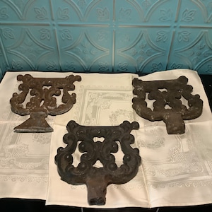 May include: Three antique cast iron architectural elements with ornate scrollwork designs. The pieces are dark gray and rest on a white cloth with a floral pattern. The background features a light blue, patterned wall.