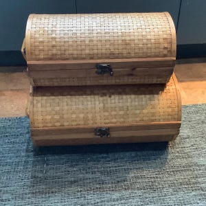 May include: Two stacked wooden storage chests with woven lids. The chests are made of natural wood with a light brown finish. The lids are woven with a light brown material.