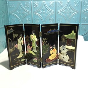 May include: A four-panel black lacquer screen with relief carvings of Asian figures and landscapes. The panels are hinged and feature gold accents. The figures are dressed in traditional clothing, and the scenes include pagodas and water features.