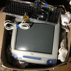 May include: A vintage iMac G3 computer in a translucent white and blue casing, featuring a built-in monitor, speakers, and a CD slot. It includes a matching translucent keyboard and a round black mouse, all resting inside a cardboard box.