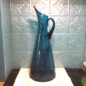 May include: A teal blue glass pitcher with a wide base and a curved spout. The pitcher has a handle on the side.