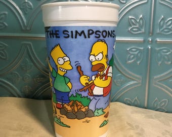 May include: A white plastic cup with a blue, yellow, and orange cartoon image of Bart and Homer Simpson from The Simpsons. The image shows them camping and building a fire. The text "The Simpsons" is printed on the cup.