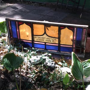 May include: A rectangular stained glass window with a wooden frame. The window features amber, blue, red, and yellow glass panels. Text is visible on the glass. The window is resting on a bed of leaves and plants.