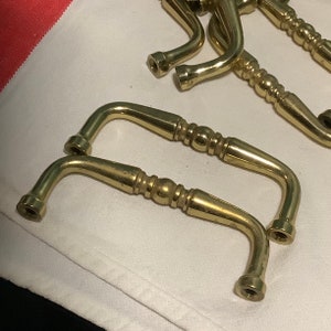 May include: Two gold-toned metal cabinet door pulls with a decorative design. The pulls are shaped like a curved 'L' with a rounded end.