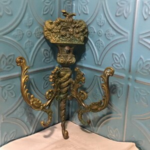 May include: Ornate brass coat hook with three hooks and a decorative top. The hook is in a vintage style and has a greenish patina.