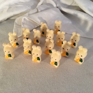 May include: A collection of small, cream-colored bunny figurines, each holding a bright orange carrot with green leaves. The bunnies have small black eyes and are arranged on a light-colored surface.