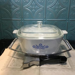 May include: White casserole dish with a blue floral design and a glass lid. The dish is sitting on a silver stand with a black handle.