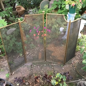 May include: A four-panel fireplace screen with a gold-colored metal frame and mesh panels. The screen has a decorative top and is set outdoors among greenery and flowers. The frame has a rope-like design along the edges.